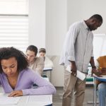 Grupo diverso de estudantes faz uma prova em uma sala de aula moderna enquanto um professor acompanha a avaliação.