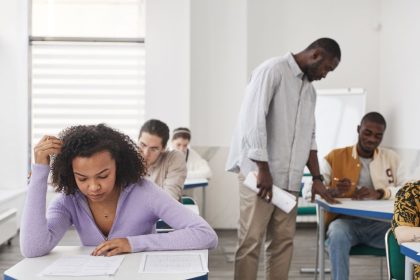 Grupo diverso de estudantes faz uma prova em uma sala de aula moderna enquanto um professor acompanha a avaliação.
