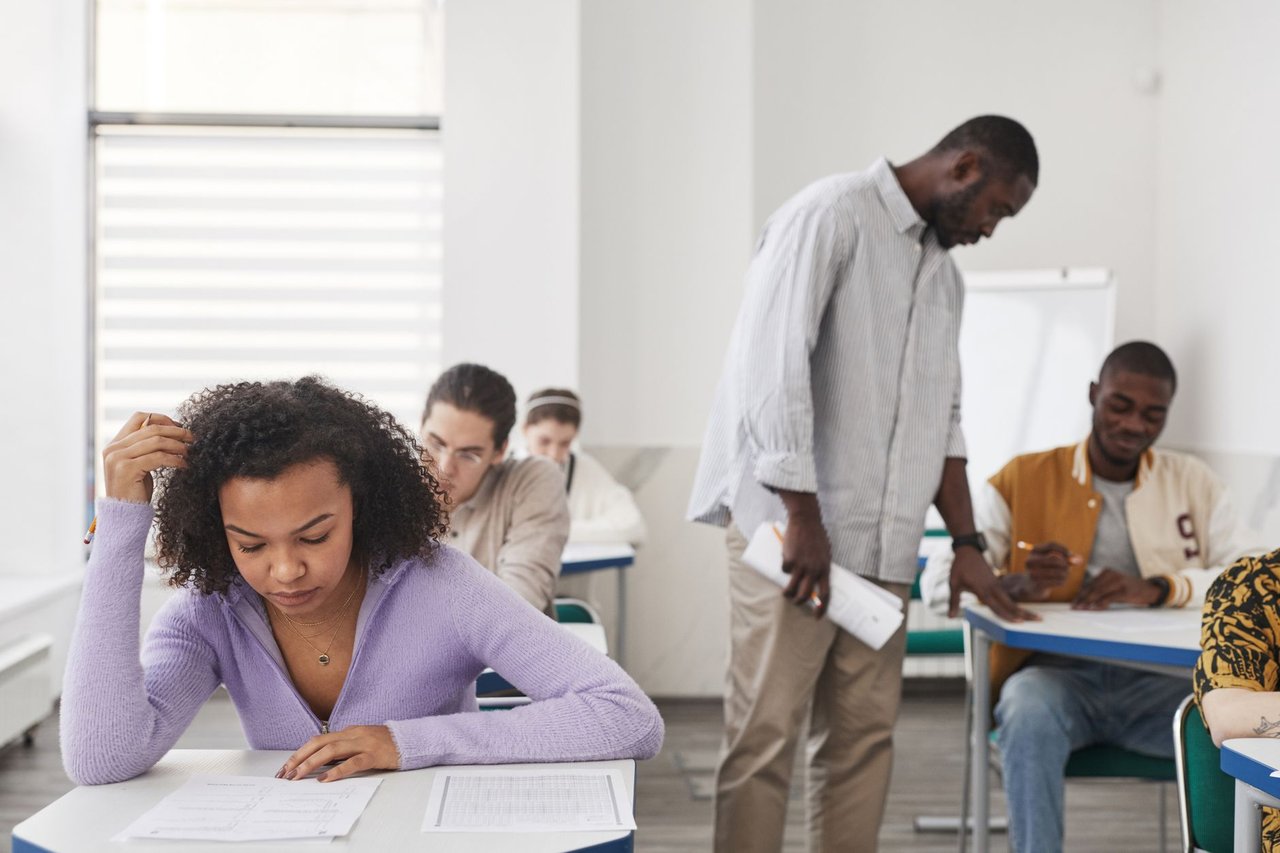 Grupo diverso de estudantes faz uma prova em uma sala de aula moderna enquanto um professor acompanha a avaliação.