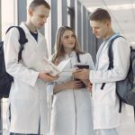 Três estudantes de medicina conversam e consultam anotações em um corredor hospitalar.