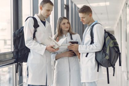 Três estudantes de medicina conversam e consultam anotações em um corredor hospitalar.