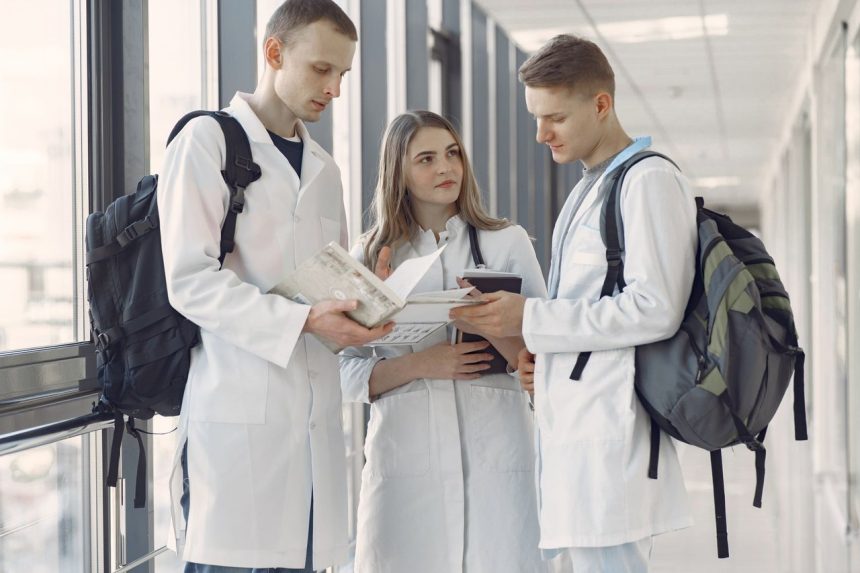 Três estudantes de medicina conversam e consultam anotações em um corredor hospitalar.