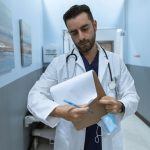 Médico de jaleco analisa um prontuário em um corredor hospitalar.