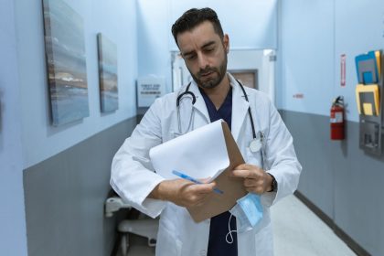 Médico de jaleco analisa um prontuário em um corredor hospitalar.