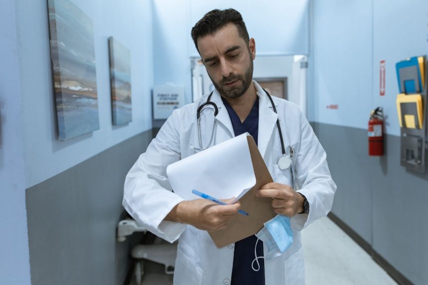 Médico de jaleco analisa um prontuário em um corredor hospitalar.