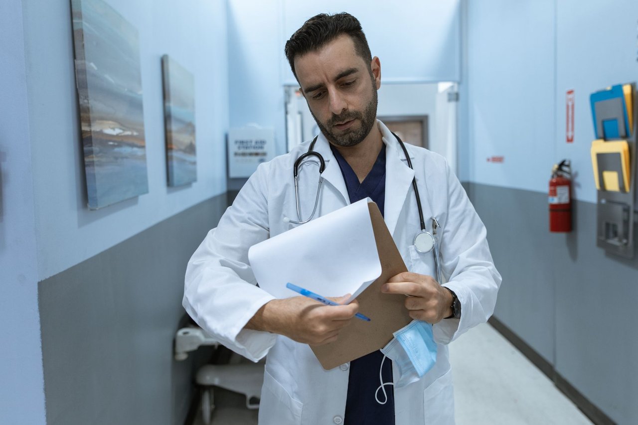 Médico de jaleco analisa um prontuário em um corredor hospitalar.