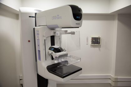 Equipamento de mamografia em uma sala hospitalar, com profissionais de saúde ao fundo.