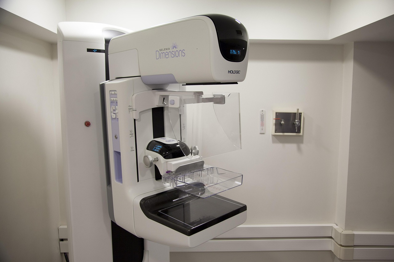 Equipamento de mamografia em uma sala hospitalar, com profissionais de saúde ao fundo.